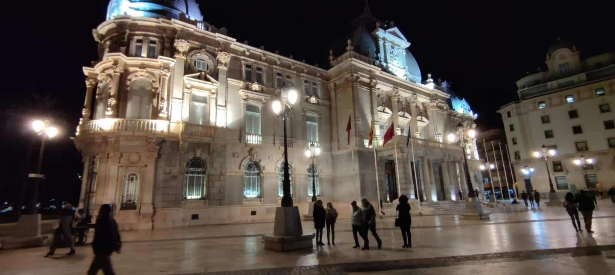Vista nocturna del Ayuntamiento de Cartagena iluminado, con transeúntes disfrutando del ambiente urbano.